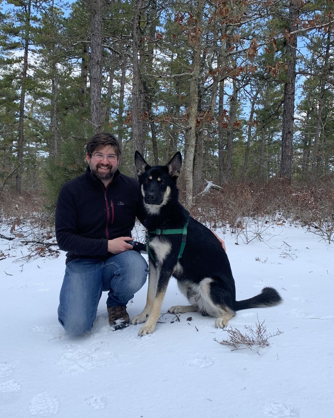 Landon, the owner of Pups On Passyunk, kneeling in the snow beside a black and tan shepherd in a green harness